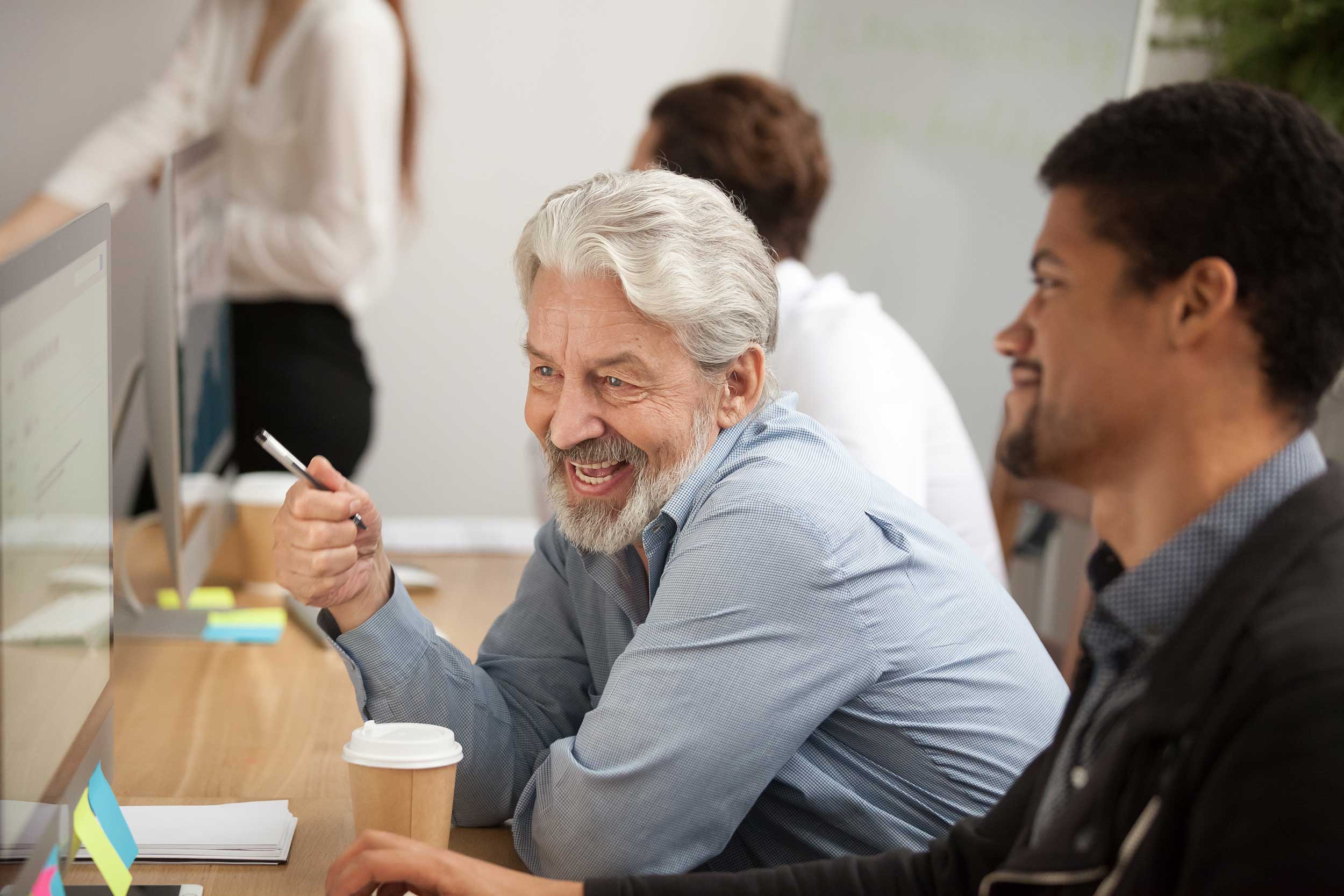 Older White Man Laughing With Younger Black Man At Desk With Takeaway Coffee